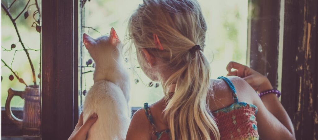 Meisje zit bij een raam en kijkt samen met haar kat naar buiten.